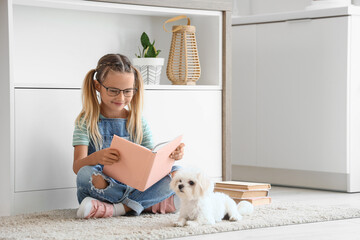 Naklejka premium Cute little happy girl with Maltese dog reading books on floor in kitchen
