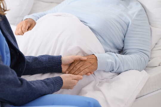 Caregiver with lying senior woman holding hands in bedroom, closeup. Care concept