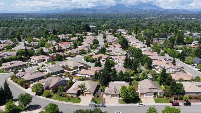 Sprawling suburban neighborhood with tile-roofed homes, lush green yards and palm trees spreads toward forested mountains under blue skies in Redding, California, USA. Aerial drone backwards rising.