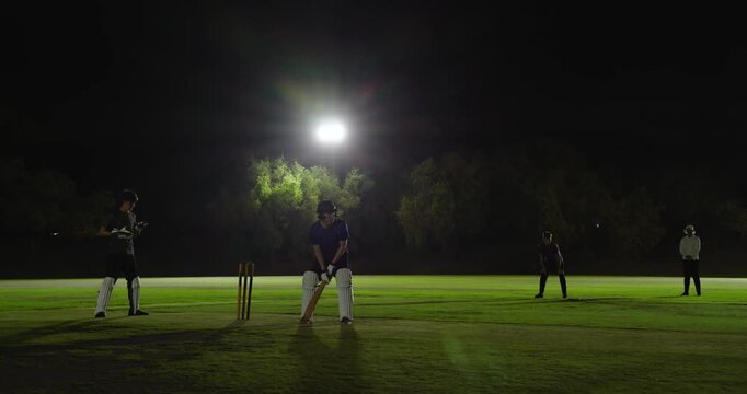 Man swinging bat on bowler's delivery under floodlight, sending ball to boundary,, hugging bowler