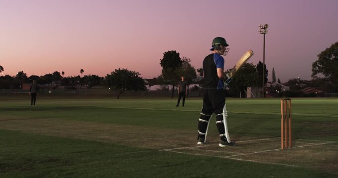 White teenage batsman batting at ground, bowler delivering and crashing stumps