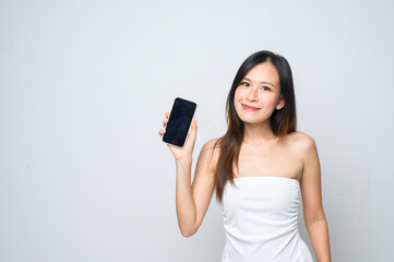 Naklejka premium A young woman in a white dress smiles while holding up a smartphone against a plain white background.