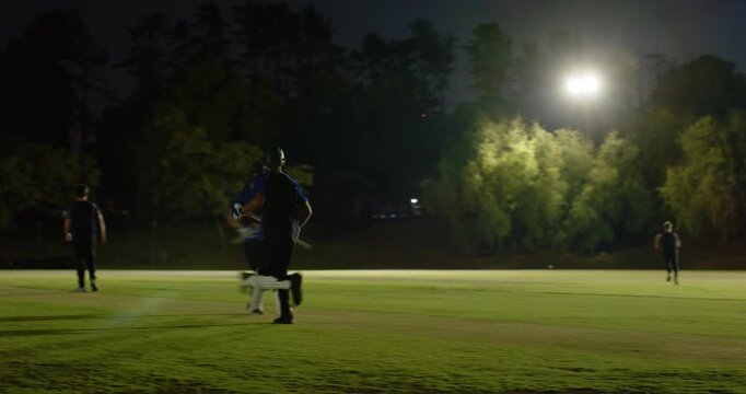 Teenage male players batting ball off bowler delivery calling teamate, sprinting under floodlights
