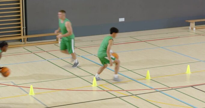 Diverse male trainees in green dribbling zig-zag through cones before overhead shot in the gym