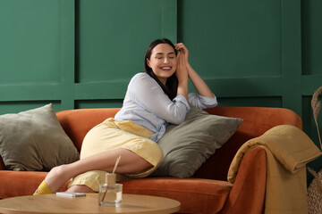 Obraz premium Young woman with cushions sitting on red sofa near green wall at home