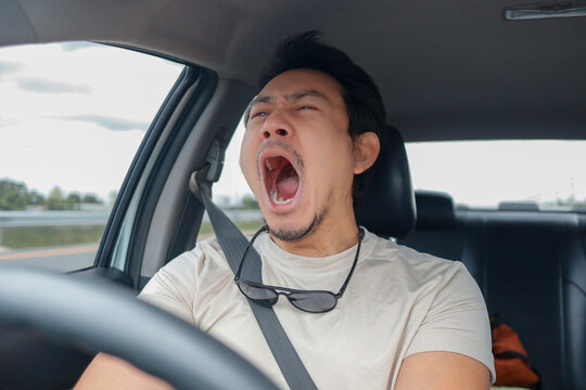 Tired Asian man yawning widely while driving a car, showing extreme fatigue and sleepiness.