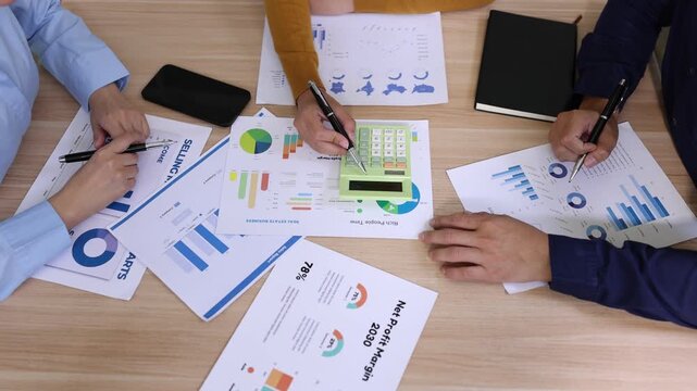 Top view of a business team analyzing financial growth charts, using a calculator and pointing at data sheets during a marketing strategy meeting on a wooden office table.