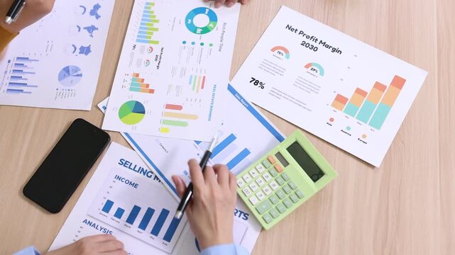 Top view of a business team analyzing financial growth charts, using a calculator and pointing at data sheets during a marketing strategy meeting on a wooden office table.