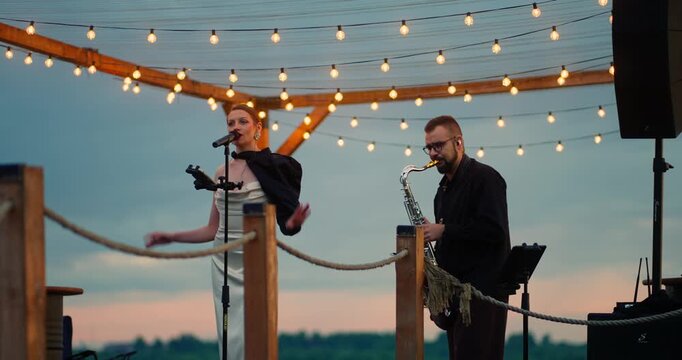 Female singer and male saxophonist performing on outdoor wooden deck at dusk