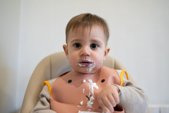 Adorable baby eating yogurt with a messy face and bib indoors