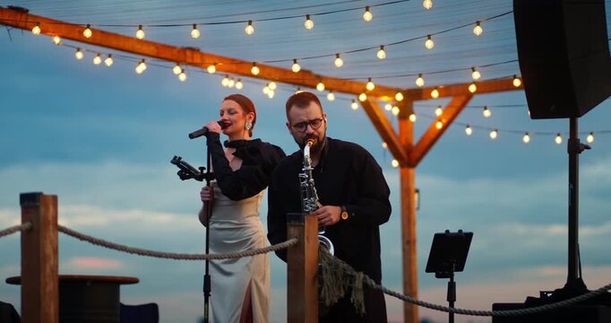 Female singer and saxophonist performing on decorated outdoor wooden deck at dusk