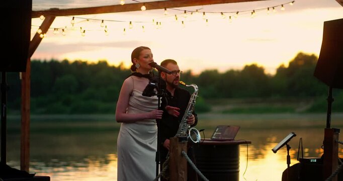Female vocalist and tenor saxophonist performing on wooden lakeside stage at sunset