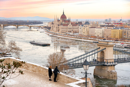 Tourists looking at winter panorama of Budapest with Hungarian Parliament and Chain Bridge over frozen Danube river at sunset, Hungary