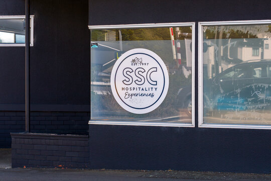 A circular hospitality‑brand logo is displayed on a storefront window, reflecting nearby buildings and street activity in New Zealand