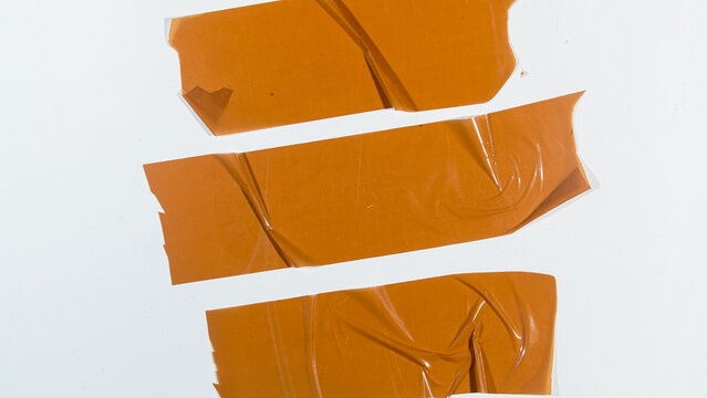 Three strips of orange adhesive tape lie on a white surface. Each strip shows creases and slight wear from use. The tape is not neatly aligned, adding a casual, lived-in feel
