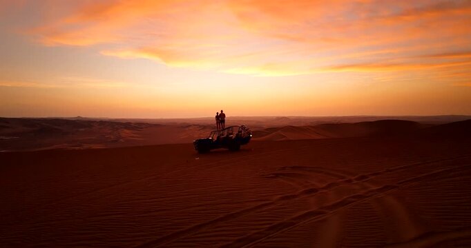 Silhouette of couple standing on roof of dune buggy watching sunset over vast sand dunes in Huacachina desert in Peru. Aerial towards, copy space