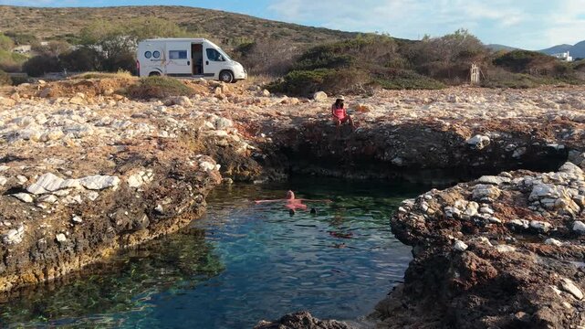 Experience the serene charm of Paros Island, Greece, where clear waters meet rocky shores. A couple relaxes by the ocean with a camper van motorhome in the background