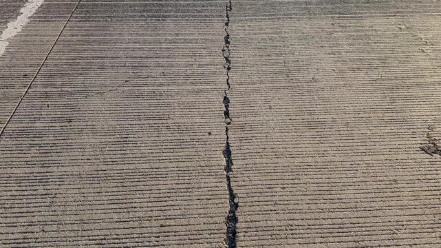 Closeup view of a cracked concrete highway surface featuring a prominent longitudinal fracture, textured pavement grooves, and signs of structural wear, captured in natural daylight from above.