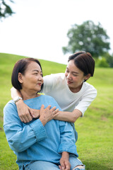 Fototapeta premium Asian adult daughter smiles and hugs her senior mother from behind in a beautiful green park. This heartwarming image captures family love, care, and joyful togetherness. Vertical Shot