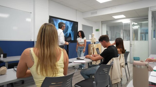 Male anatomy professor teaching a lesson to university medical students in a classroom, pointing at an x ray image of a hip joint on a large screen while a young woman assists