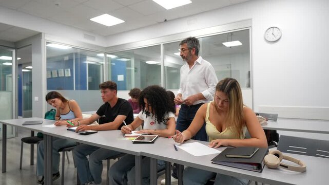 University teacher walking through the classroom and monitoring students during a lesson or examination. Diverse group of young people focused on writing their test at their desks in a college hall
