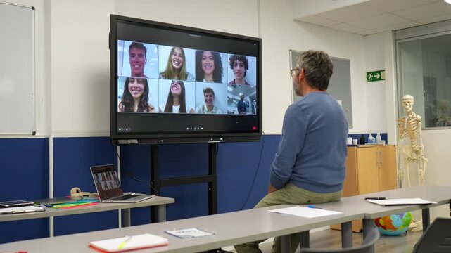 Middle aged male teacher sitting on a desk in a modern classroom, having a video conference with his students on a large digital screen during a hybrid lesson for remote learning