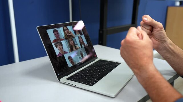 Over the shoulder view of a professor talking and gesturing during a video call on a laptop with a group of young students in a grid view, representing remote education and e learning concepts
