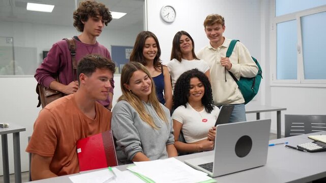 Cheerful multiracial group of high school students gathered around a laptop, smiling and waving during a video conference call, collaborating on a project together in a modern classroom