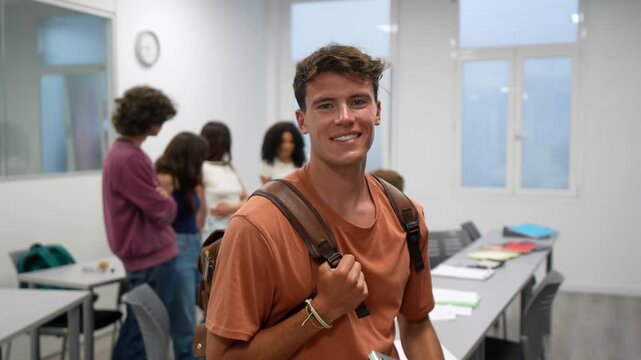 Portrait of a happy young male student with a backpack and a laptop smiling at the camera inside a modern university classroom, with other classmates talking in the background