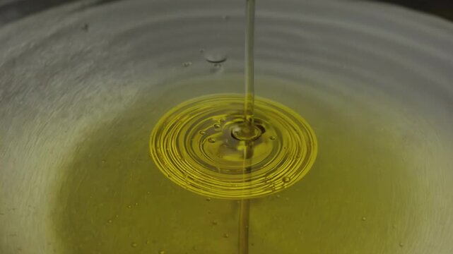 Liquid oil is poured into a shallow metallic bowl, creating ripples and circular patterns on the surface as it flows and interacts with the water below