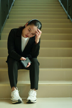 Young asian businesswoman sitting on office stairs, feeling stressed and suffering from burnout, holding a small empty wallet, symbolizing job loss, unemployment, or financial hardship