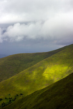 Green rolling mountain hills illuminated sunlight beneath dramatic low clouds overcast sky.
