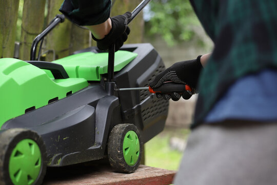 Men's hands in work gloves repair a lawn mower with a screwdriver in the yard.