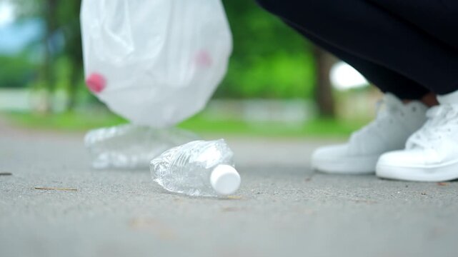 Close-up hand in a blue glove picking up a plastic bottle from a park path. Blurred volunteer in background. Concept of environmental conservation, plogging, and recycling.