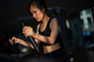 Fototapeta premium A focused woman in black workout attire uses an elliptical machine in a dimly lit gym.