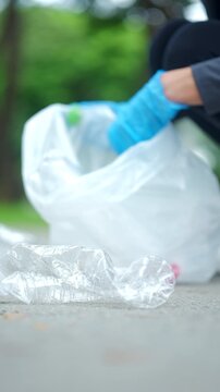 Close-up hand in a blue glove picking up a plastic bottle from a park path. Blurred volunteer in background. Concept of environmental conservation, plogging, and recycling.