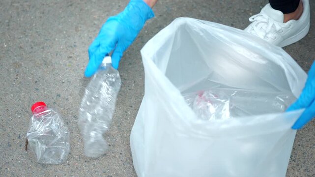 Close-up hand in a blue glove picking up a plastic bottle from a park path. Blurred volunteer in background. Concept of environmental conservation, plogging, and recycling.