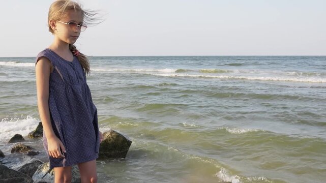 A girl in a blue dress with polka dots and sunglasses looks into the distance on the beach