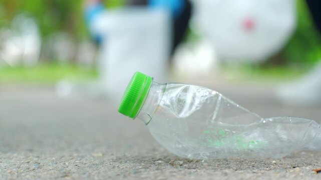 Close-up hand in a blue glove picking up a plastic bottle from a park path. Blurred volunteer in background. Concept of environmental conservation, plogging, and recycling.