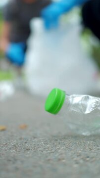 Close-up hand in a blue glove picking up a plastic bottle from a park path. Blurred volunteer in background. Concept of environmental conservation, plogging, and recycling.