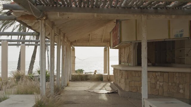 Empty ruined beach bar in old hotel resort by the sea during sunset or sunrise. Neglected property or foreclosure rundown business. Swimming pool bar closed and broken with dry palm tree leaves roof
