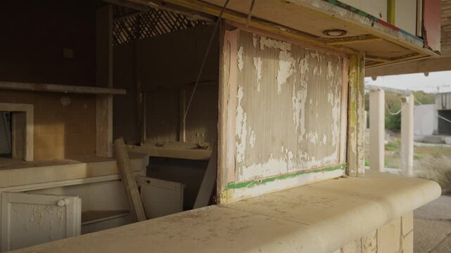 Abandoned stall with peeling paint and broken counter. Sunlight falls through open doorway. Inside, shelves are empty and dusty. Structure shows signs of neglect and time