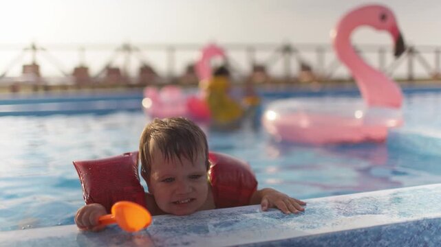 A boy with armbands and a disgruntled face, trying to get out of a pool of clean water and calling for his mother