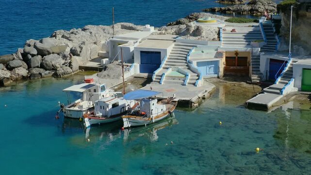 Glistening turquoise waters frame a quaint harbor in Mandrakia Milos Island. Traditional boats sway gently, while picturesque steps lead to charming coastal buildings under a clear blue sky