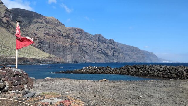 Scenic wide angle view of massive ocean cliffs and volcanic stone shore with a red warning flag waving in the wind, majestic rocky coast and blue sea travel destination in Canary Islands, Spain