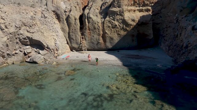 Experience the serene allure of Milos, Greece. Discover vibrant turquoise waters and dramatic cliffs as visitors enjoy a sun-soaked day on the Tsigrado Beach