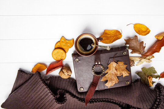 Cup coffee with book in leather binding autumn dry leaf and warm scarf on white wooden board top view.