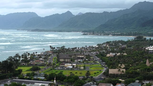 Coastal Hawaiian town with palm trees and low-rise buildings nestled between dramatic green mountain ridges and a choppy turquoise ocean shoreline in Laie, Hawaii, USA. Aerial drone wide shot
