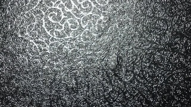 Close-up video showing a black textured surface with subtle embossed floral and ornamental glass patterns illuminated by shifting light. Ideal for abstract backgrounds, luxury dark textures, interior 