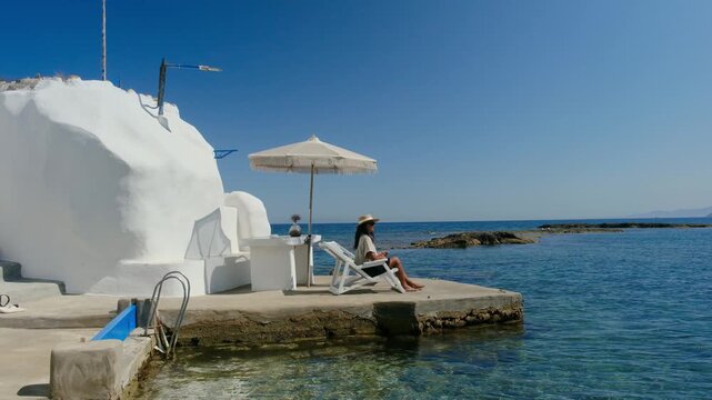 A woman enjoy a peaceful day by the azure waters of Mandrakia Milos Island, Greece. A charming white structure and umbrella create the perfect setting for leisure and reflection under the sun